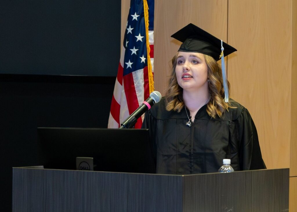 Girl in graduation gown and mortarboard standing at a podium with American flag behind her