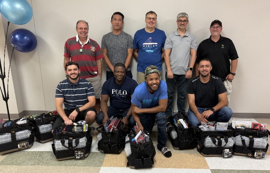 Nine adult students pose for a graduation celebration photo with their electrician's gear bags